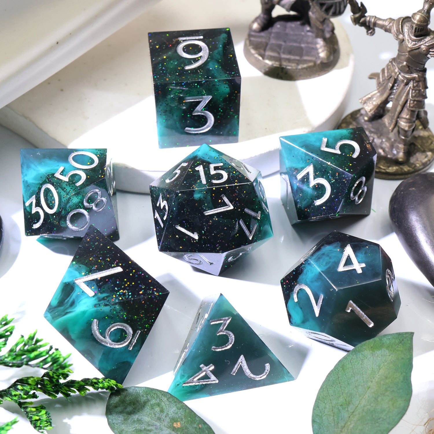 A set of teal and black glitter polyhedral gaming dice with silver numbers, arranged on a white surface with decorative leaves and figurines in the background.