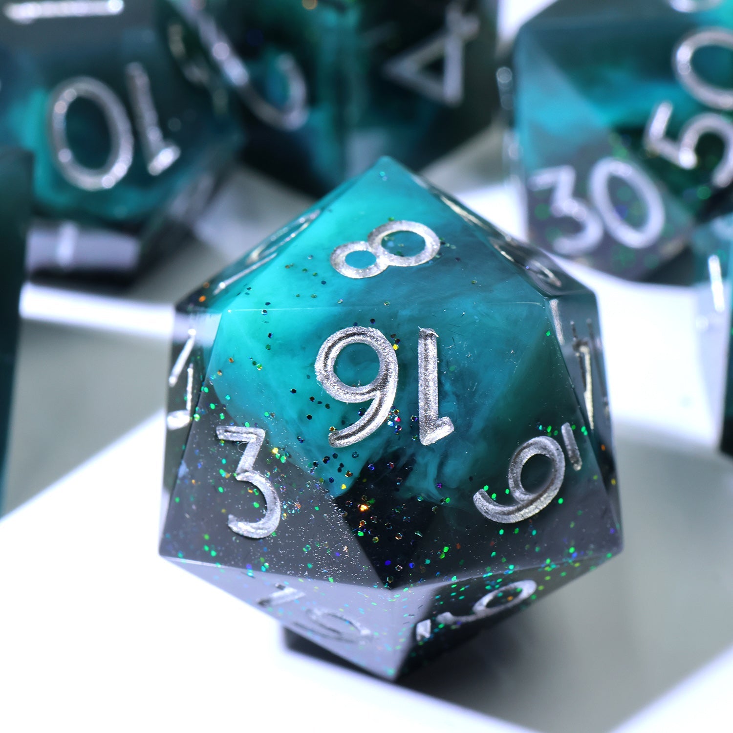 A close-up of a translucent teal and black polyhedral gaming die with silver numbering, surrounded by other matching dice on a white surface.