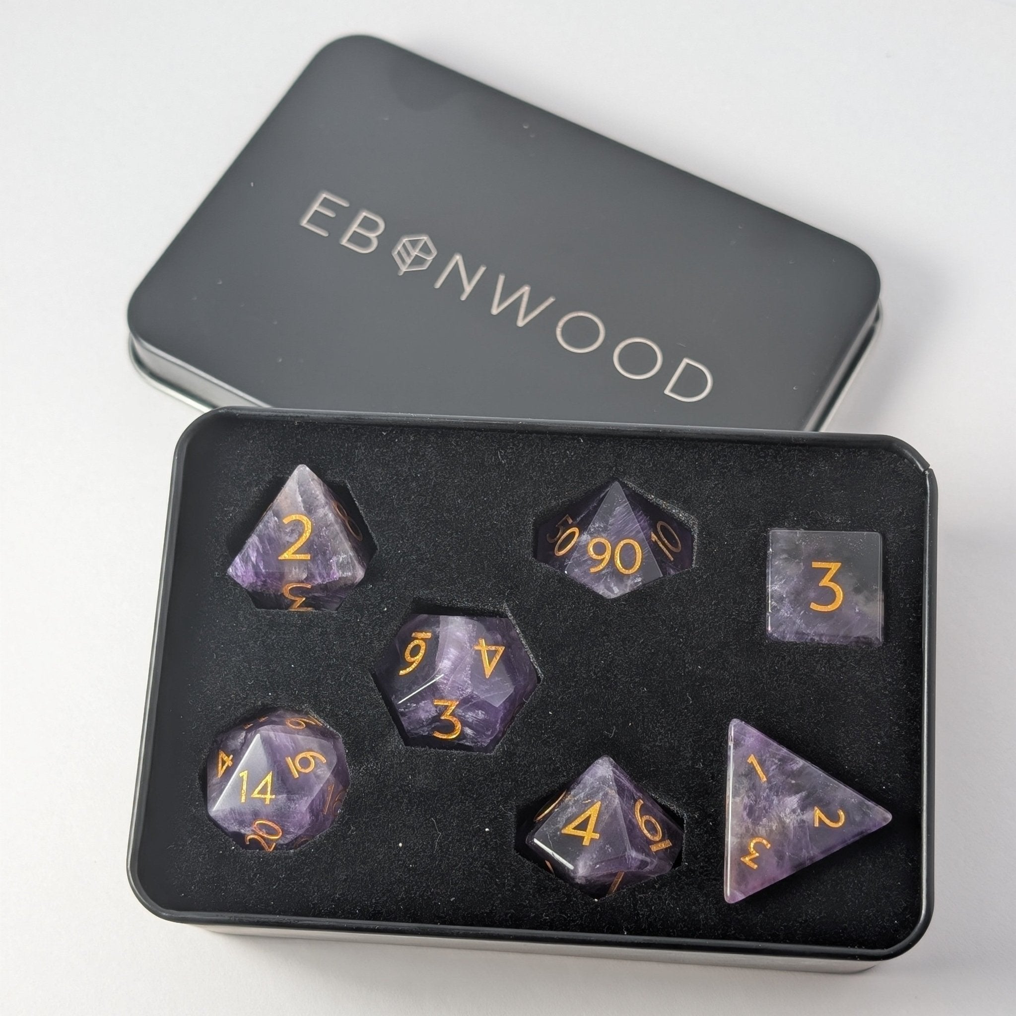 Ebonwood's Amethyst gemstone D&D dice set neatly organized within a custom metal dice storage tin, with individual compartments protecting each polyhedral die and showcasing the minimalist gold-inked font.