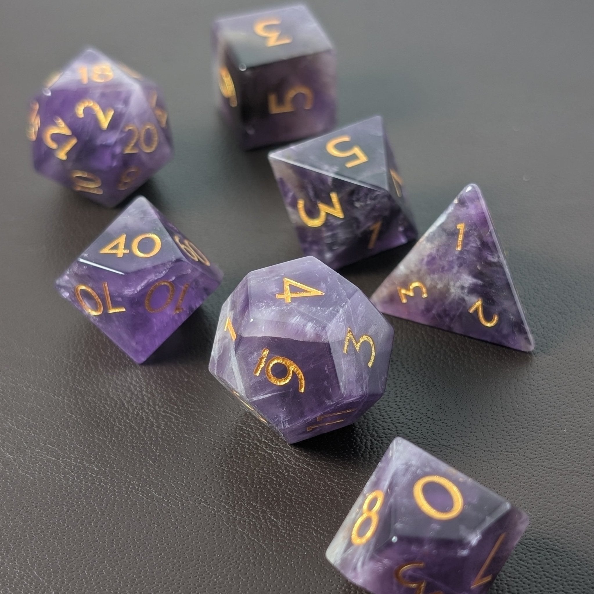 Ebonwood's full Amethyst gemstone D&D dice set with minimalist gold-inked font, resting on a premium leather dice rolling tray, emphasizing safe play and luxurious tabletop gaming.