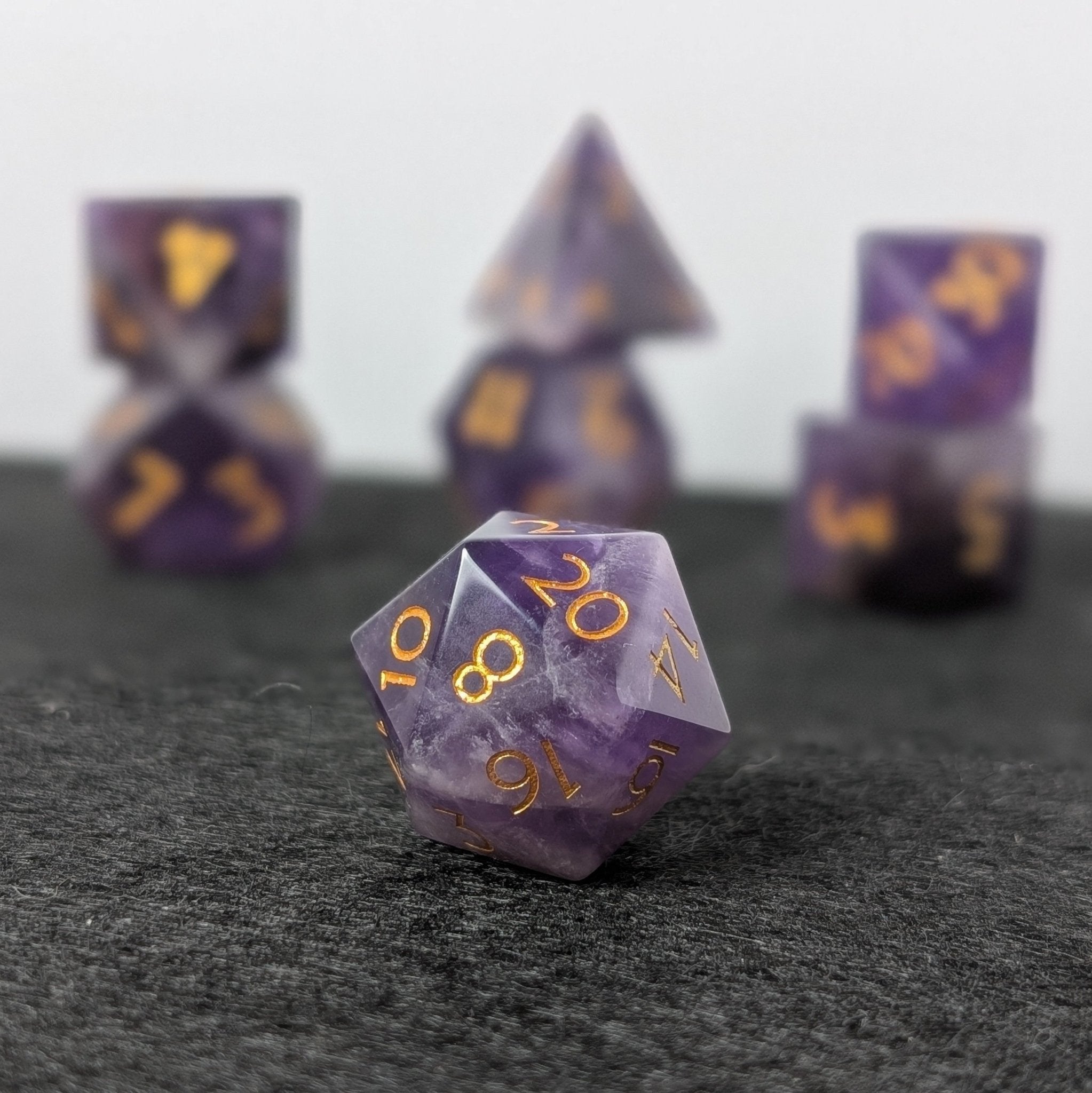 Complete 7-piece set of Ebonwood's natural Amethyst polyhedral D&D dice with the D20 prominently displayed in front, featuring rich purple tones and minimalist gold-inked numbers.