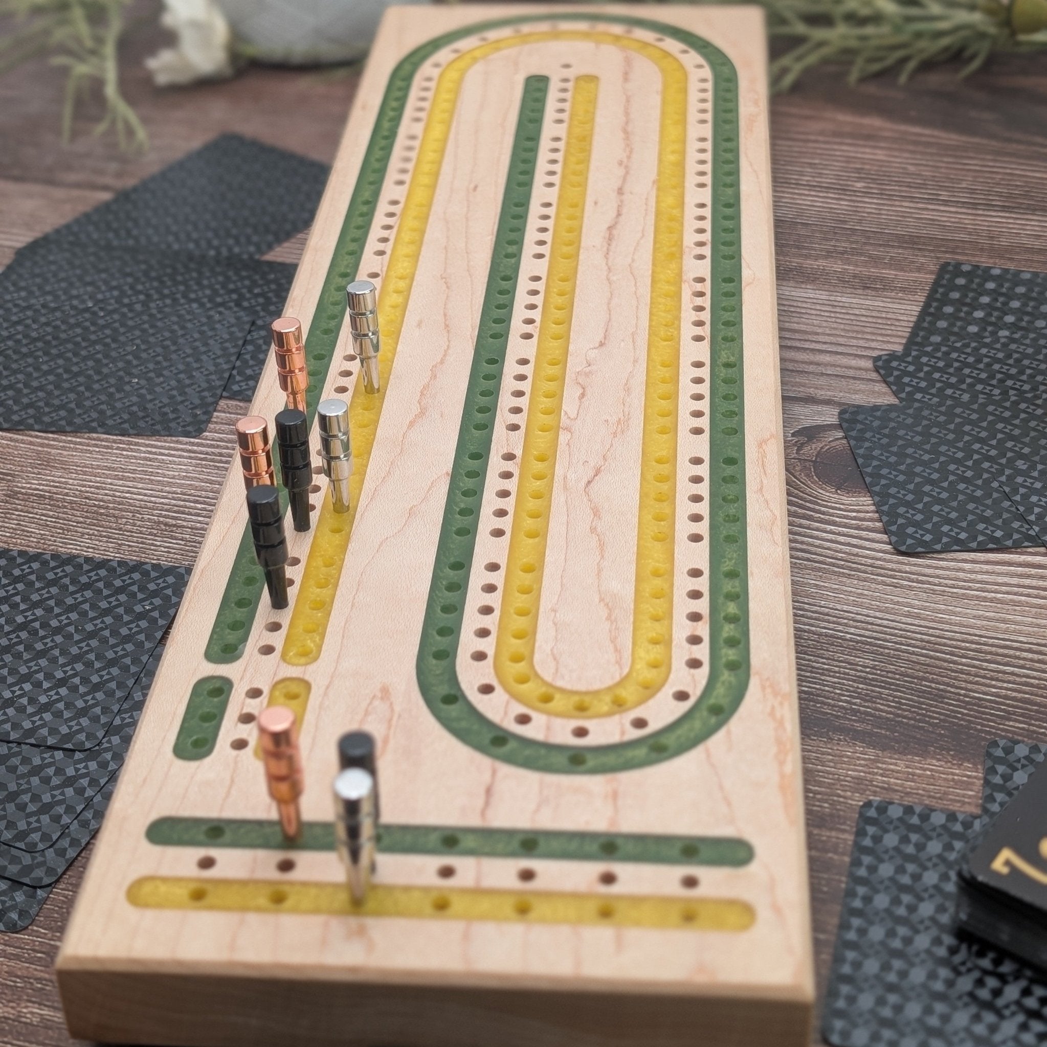close up of a green and gold cribbage board in maple wood on wood grain background with black cards on table