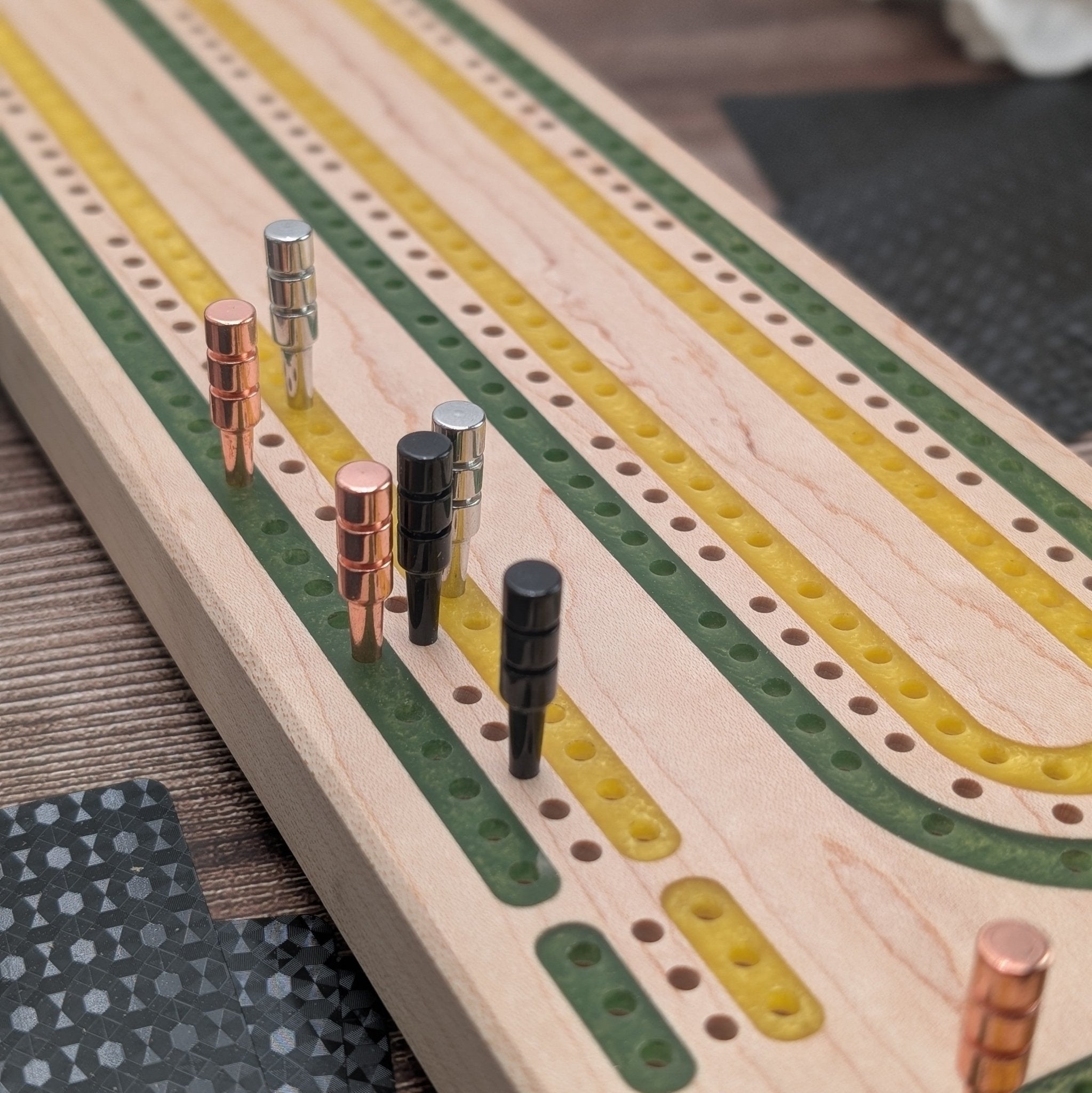 up close shot of a green and gold maple cribbage board with copper pegs in the green lane, black pegs in the wood lane, and silver pegs in gold lane