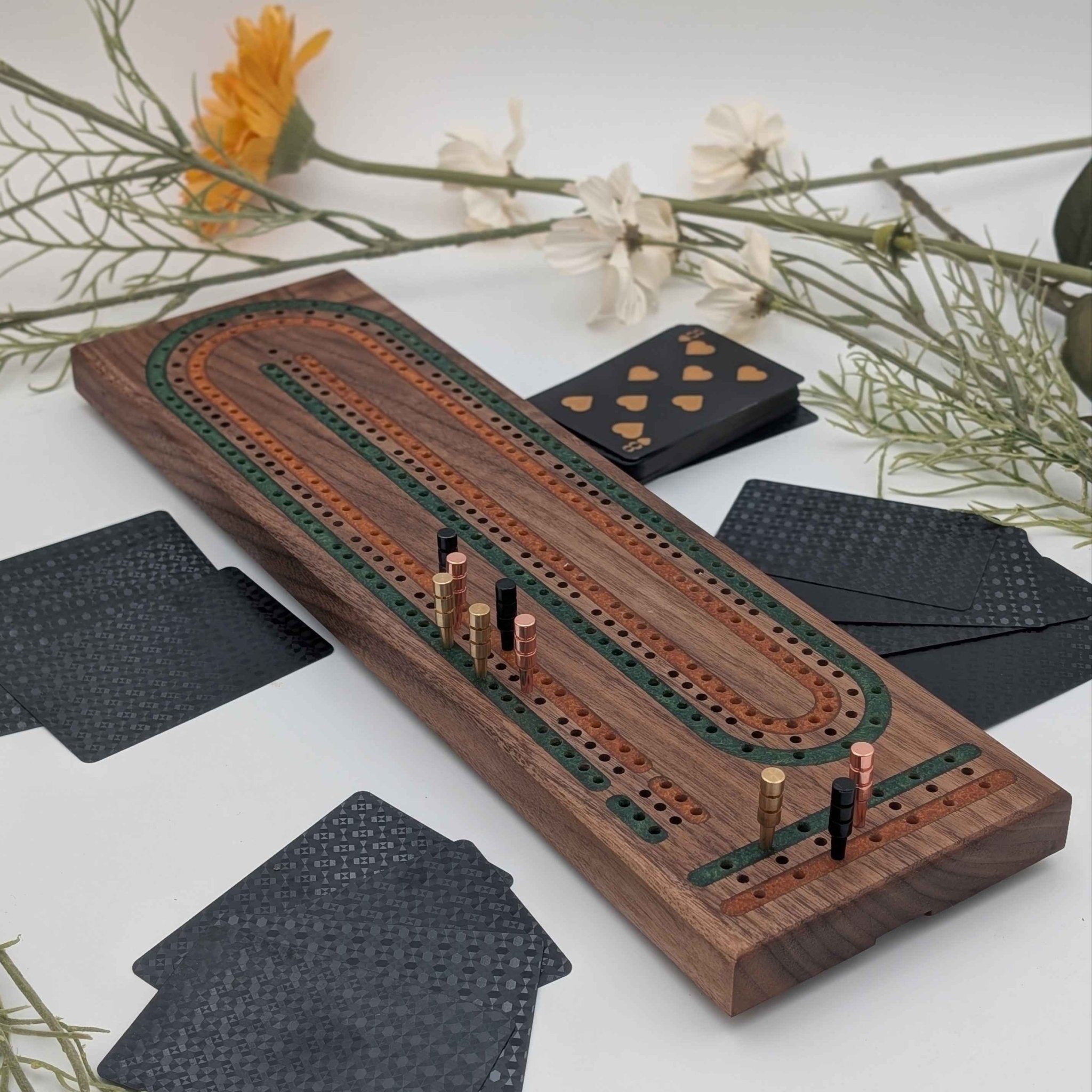 luxury cribbage board made of walnut wood with multi-color green and gold resin inlays. Photo includes black and gold cards on white background and board has gold, rose gold, and black pegs shown.