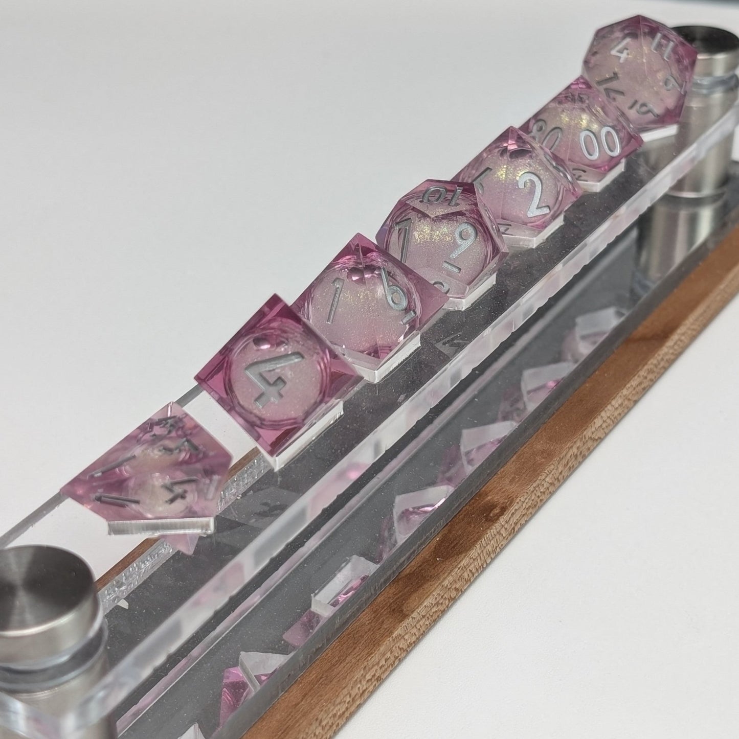 A handcrafted pink sharp-edged polyhedral DND dice set with a glitter-filled liquid core and silver numbers, made for TTRPGs and roleplaying games on top of a dice shelf with a white background