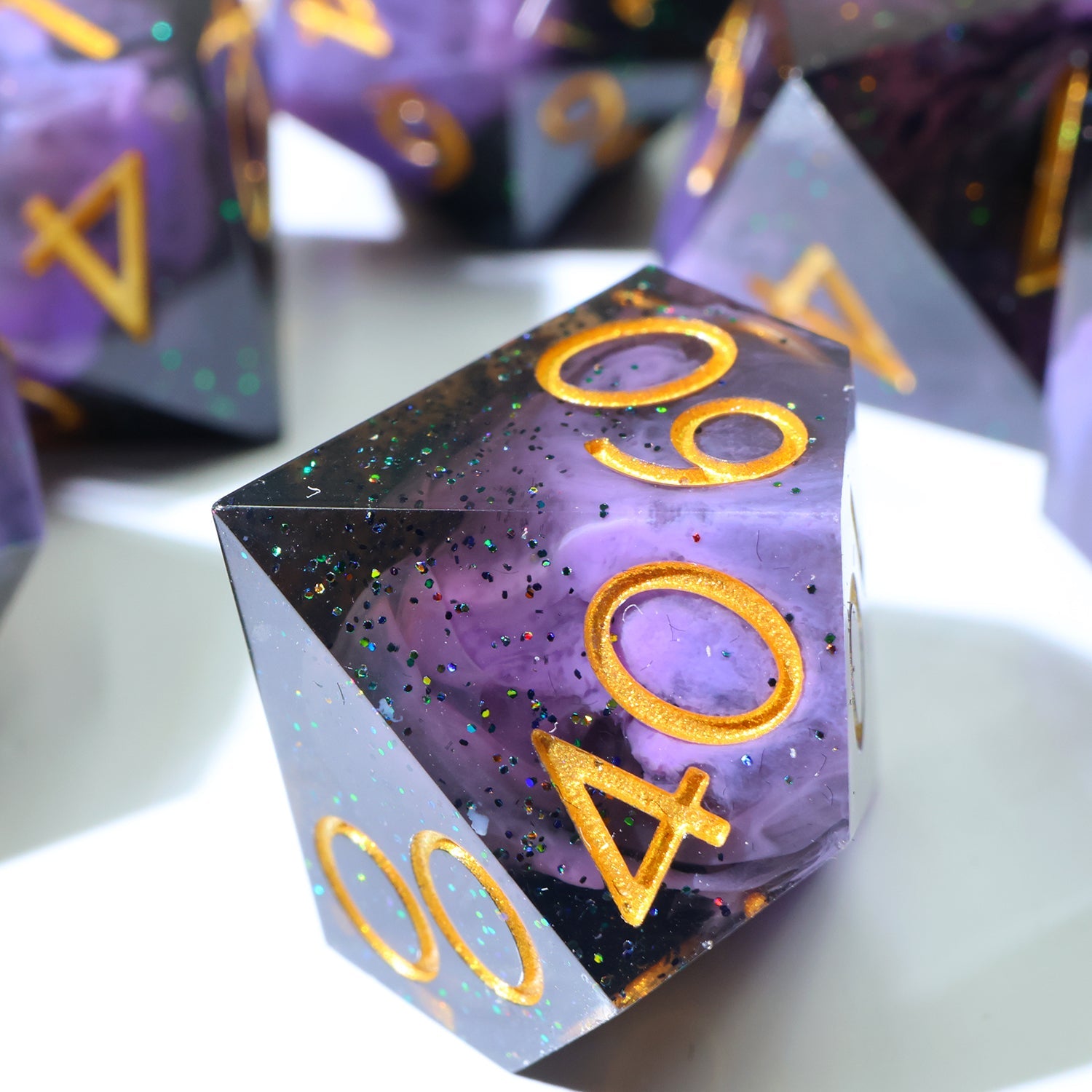 A purple and black glittery polyhedral gaming die with gold-colored numbering, shown in a close-up shot with other similar dice blurred in the background.