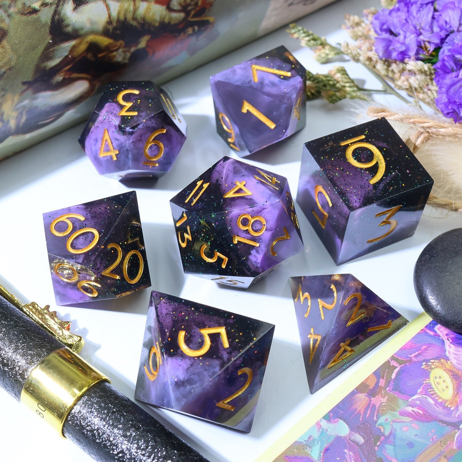 A set of purple and black polyhedral gaming dice with gold numbers, arranged on a white surface with decorative dried flowers, a black and gold pen, and a colorful card.