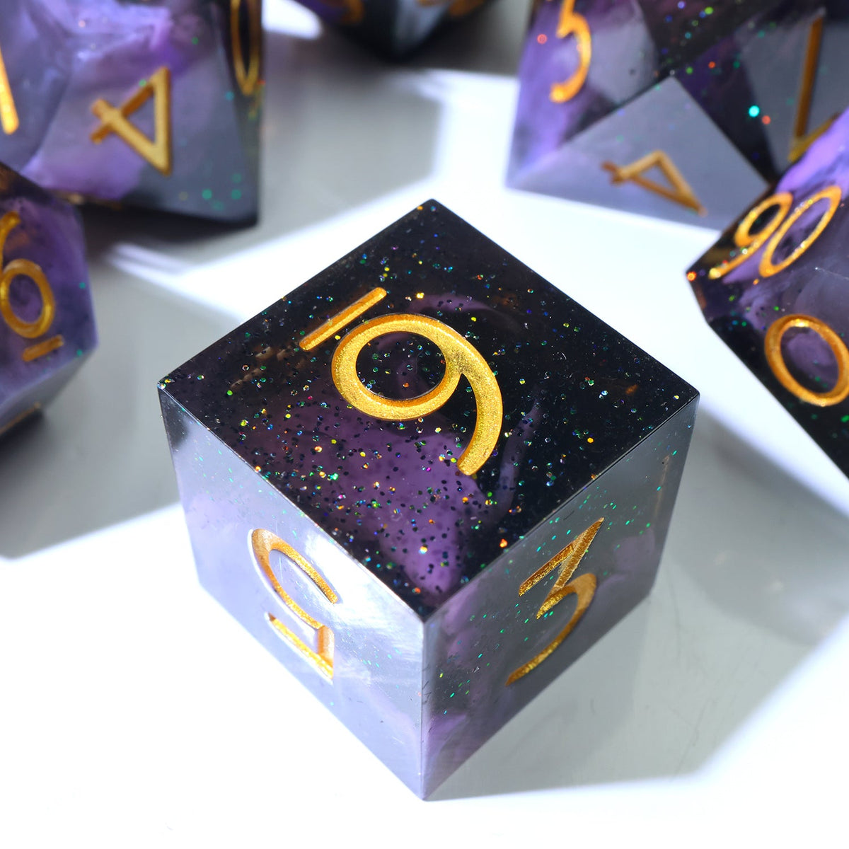 A close-up of a purple and black glittery polyhedral die, showing a gold number 19 on top, with other matching dice blurred in the background on a white surface.
