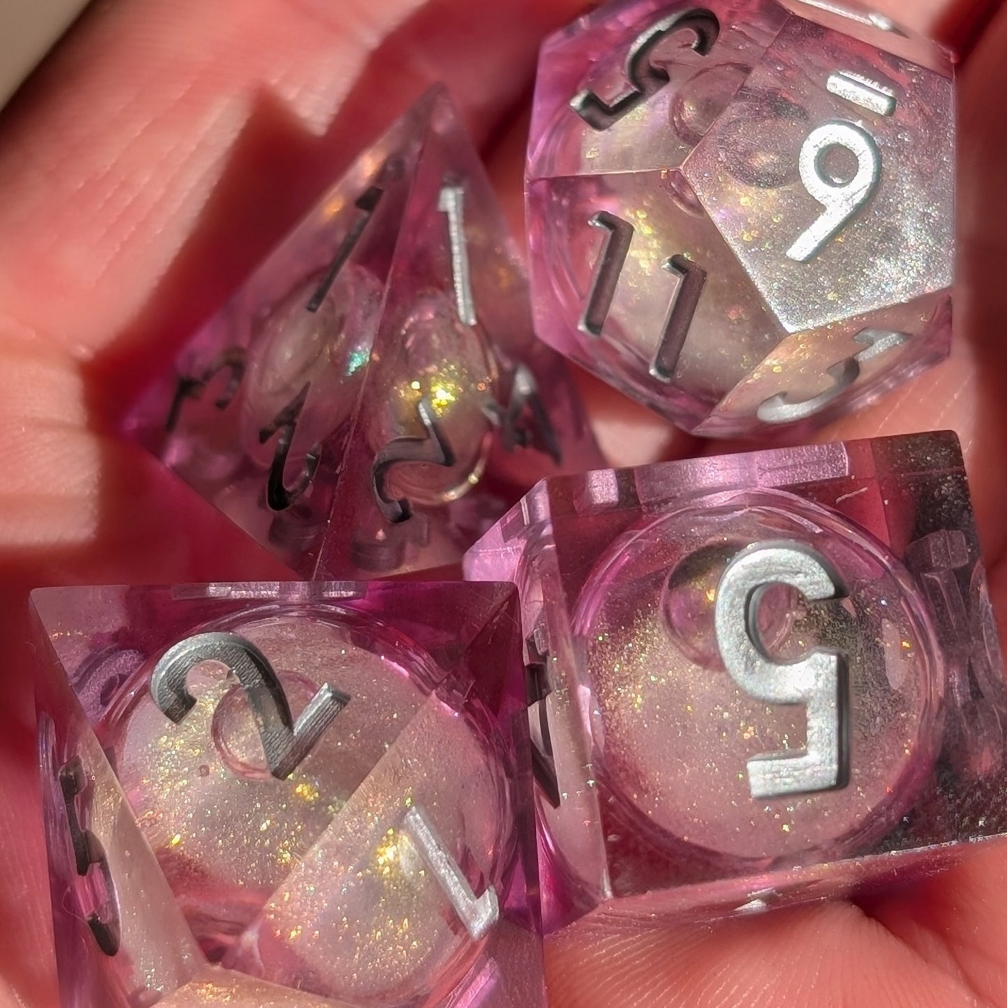 A handcrafted pink sharp-edged polyhedral DND dice set with a glitter-filled liquid core and silver numbers, made for TTRPGs and roleplaying games.