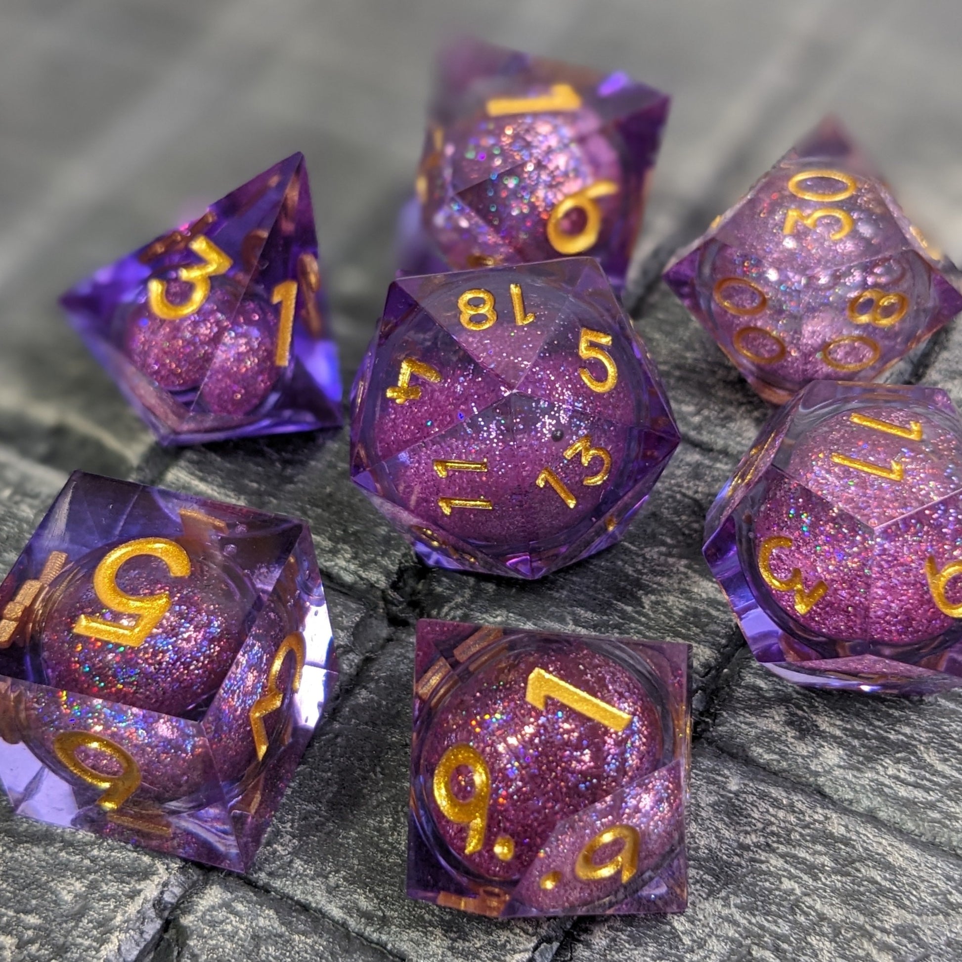 Set of 7 liquid core dice with purple resin and gold numbers on a grey stone like backdrop used in dungeons & dragons.