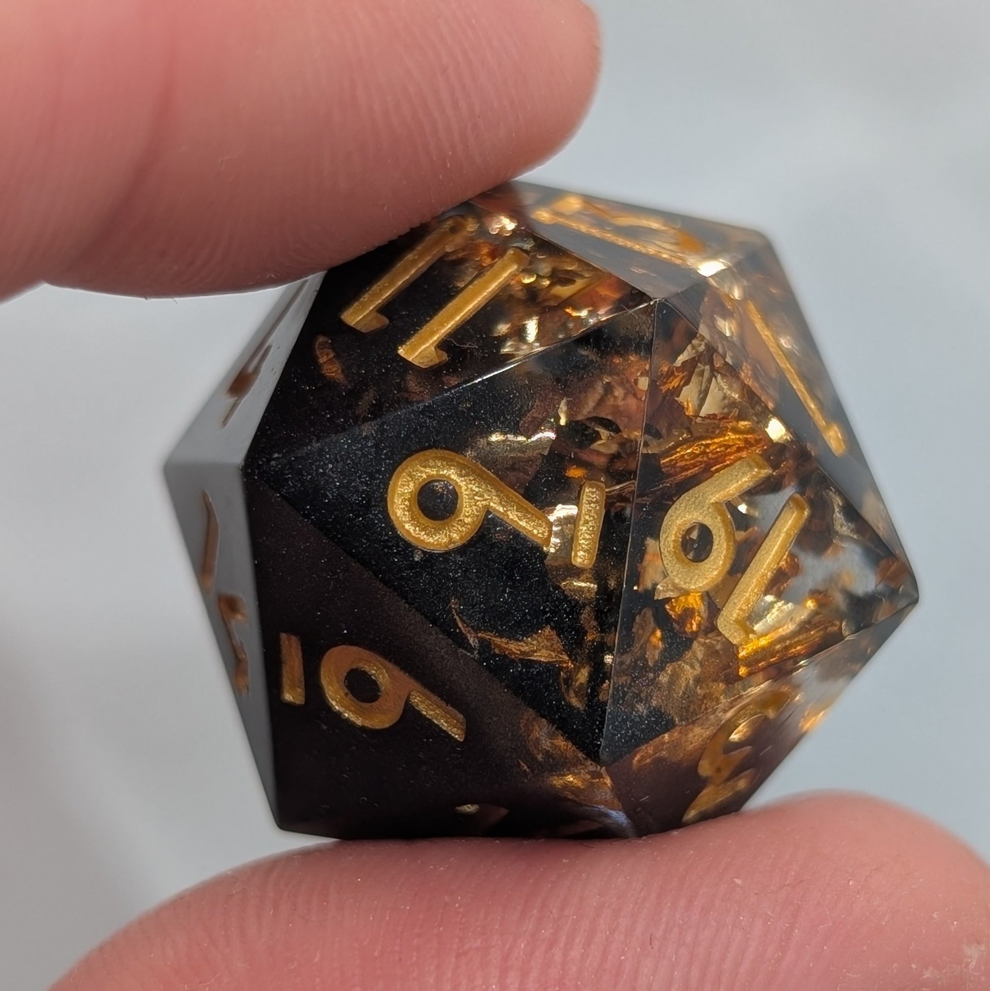 black and gold d20 with gold numbers being held between two fingers with off white background