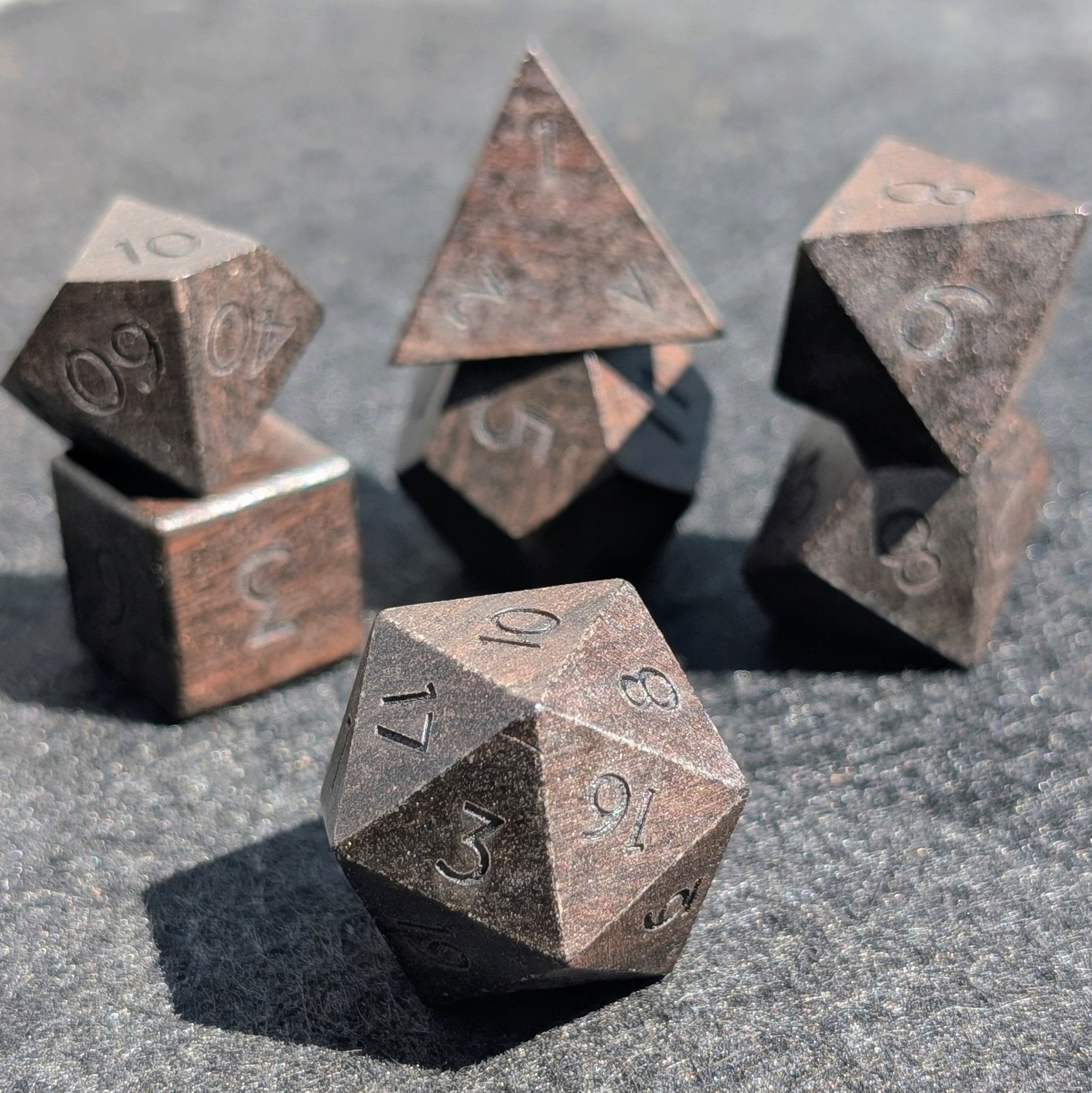 A set of polyhedral tabletop gaming dice with a weathered metallic bronze finish, arranged on a textured gray surface.
