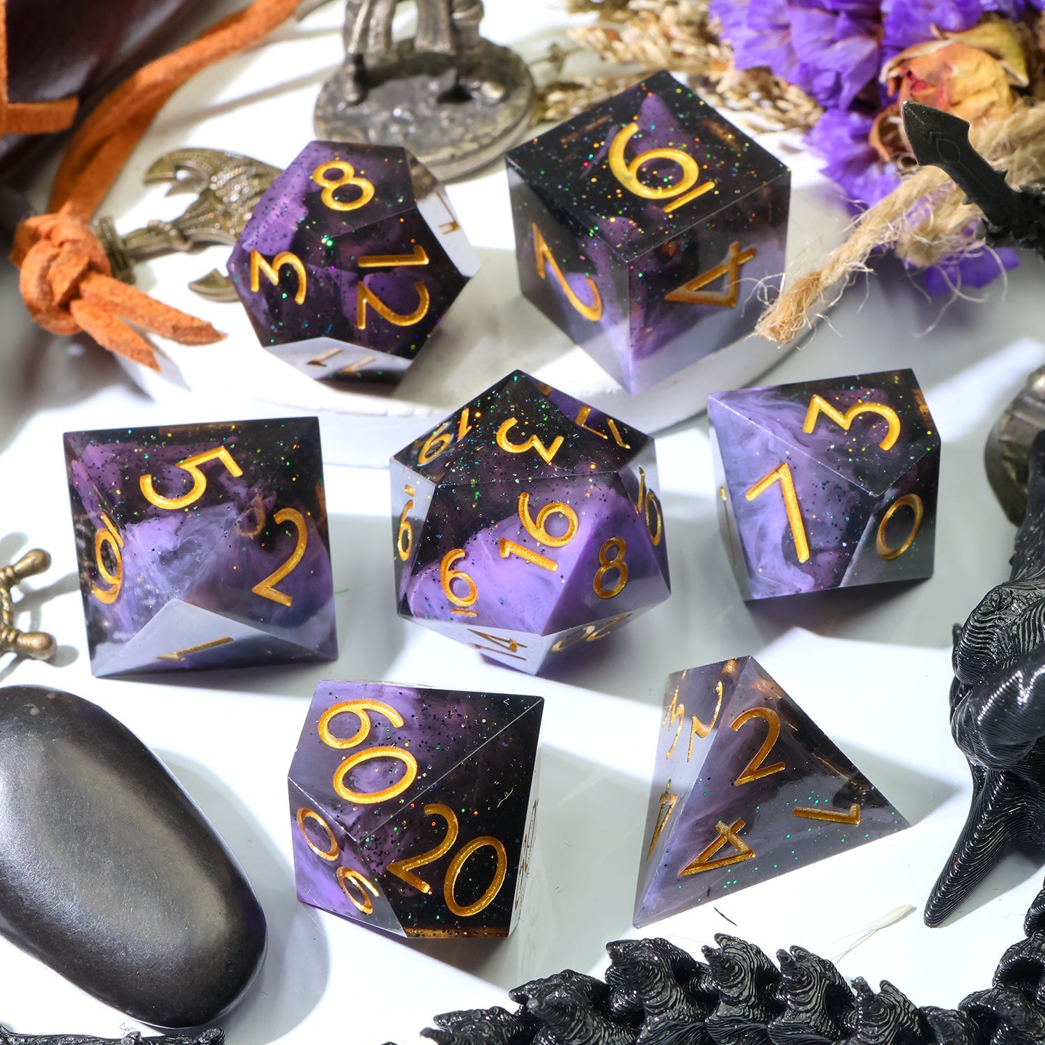 A set of purple and black polyhedral gaming dice with gold numbers, arranged on a white surface with decorative occult-themed props including a black raven figurine, dried flowers, and a metal cauldron.