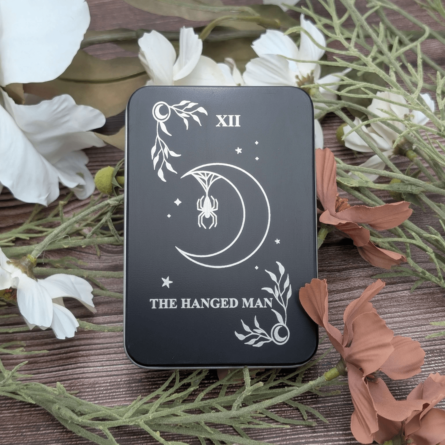 The Hanged Man tarot inspired dice tin for polyhedral dice storage on a woodgrain background with flowers. Engraving features a moon with stars and a spider hanging from the moon from its web.