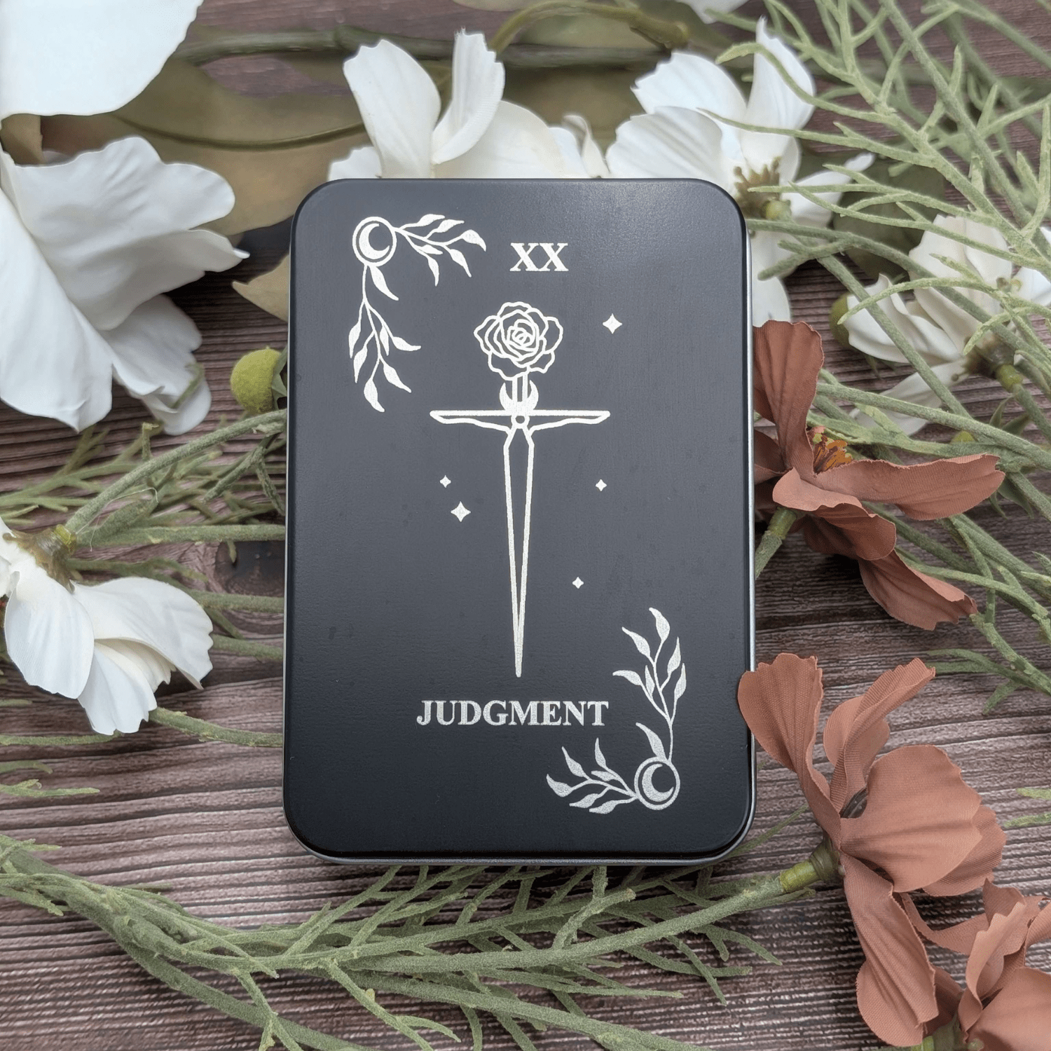 Judgement tarot inspired dice tin for polyhedral dice storage on a woodgrain background with flowers. Engraved is a celestial corners with floral dagger in center.