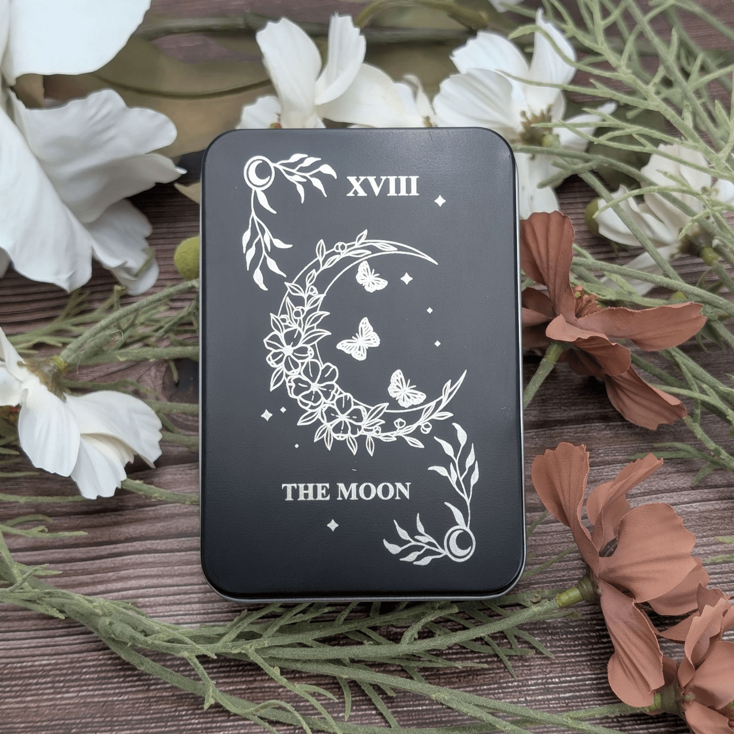 The Moon tarot inspired dice tin for polyhedral dice storage on a woodgrain background with flowers featuring a floral moon with butterflys and stars in the center.