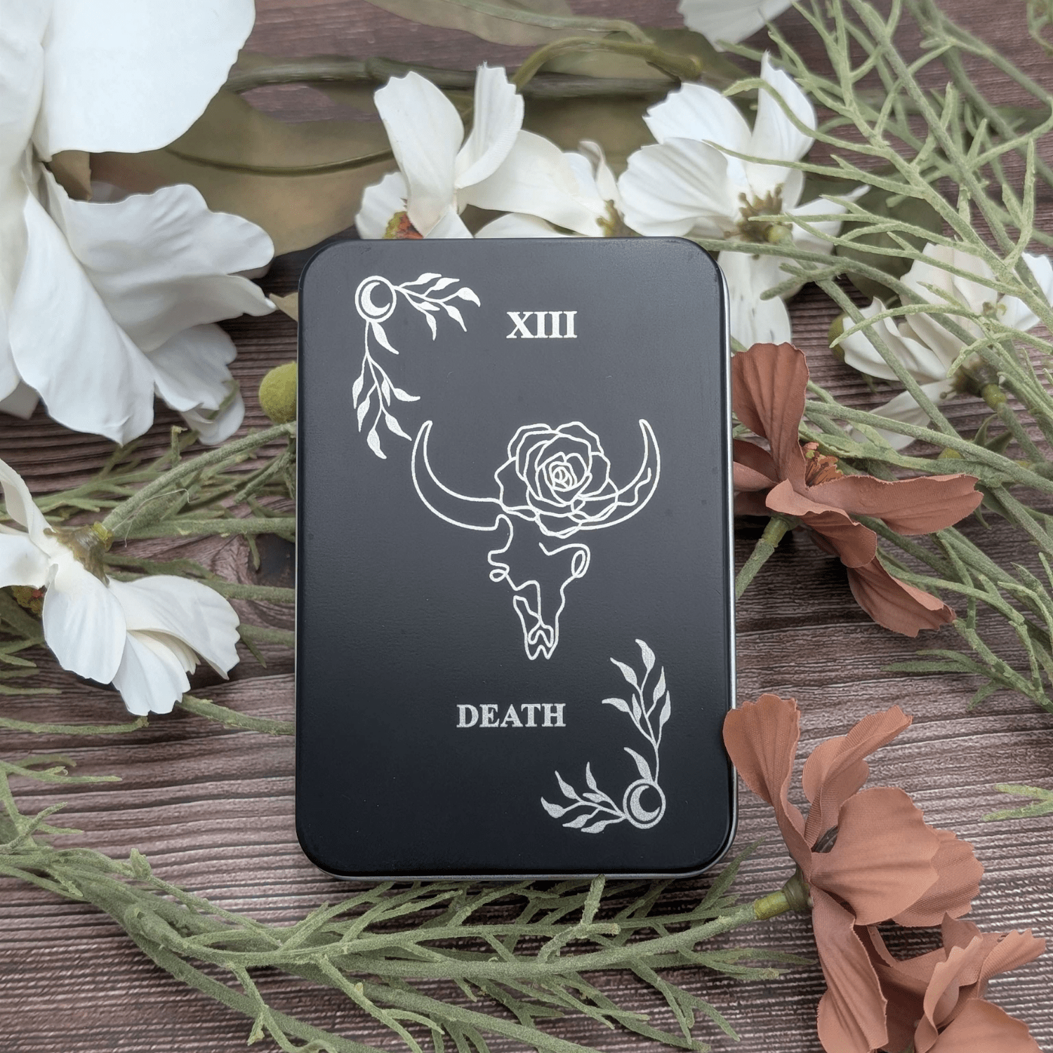 Death tarot inspired dice tin for polyhedral dice storage on a woodgrain background with flowers