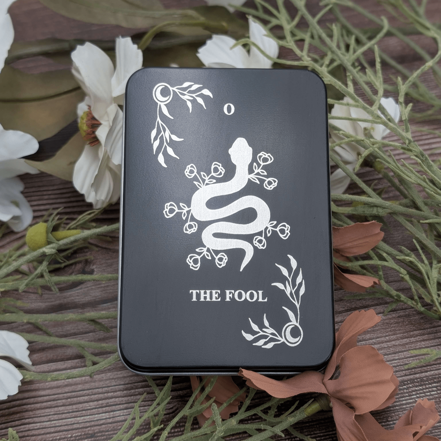 The Fool tarot inspired dice tin for polyhedral dice storage on a woodgrain background with flowers featuring a snake engraving with flowers coming out and celestial corners