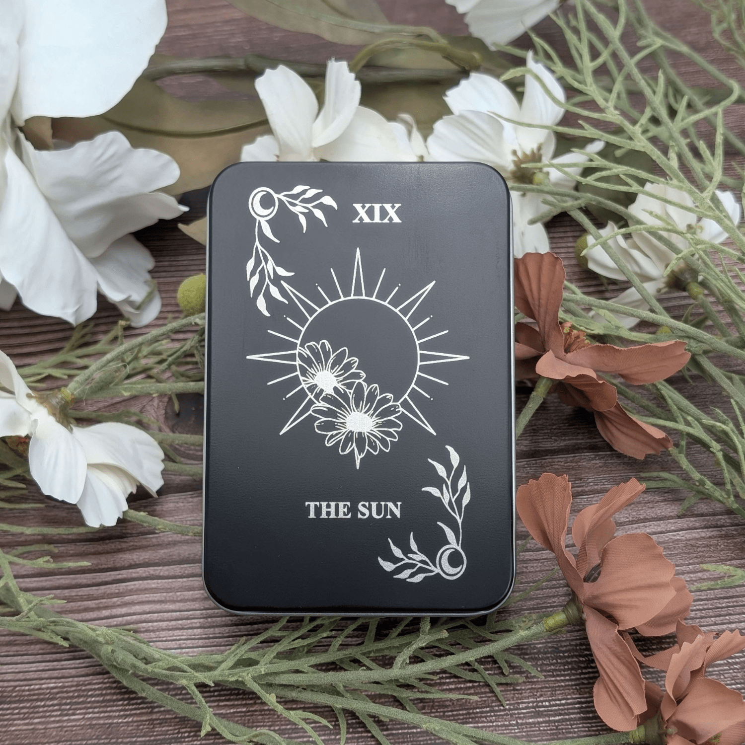 The Sun tarot inspired dice tin for polyhedral dice storage on a woodgrain background with flowers featuring a floral sun engraving with celestial corners.