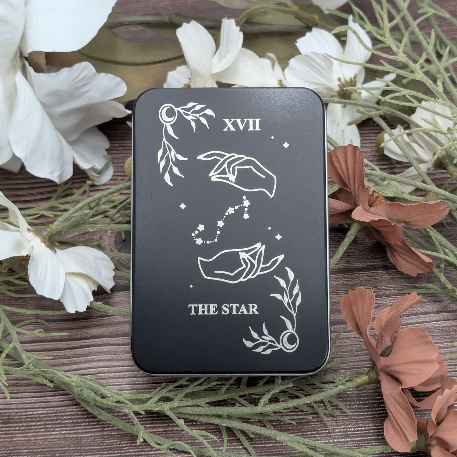 The star tarot inspired dice tin for polyhedral dice storage on a woodgrain background with flowers featuring two hands making magic stars in a celestial shape